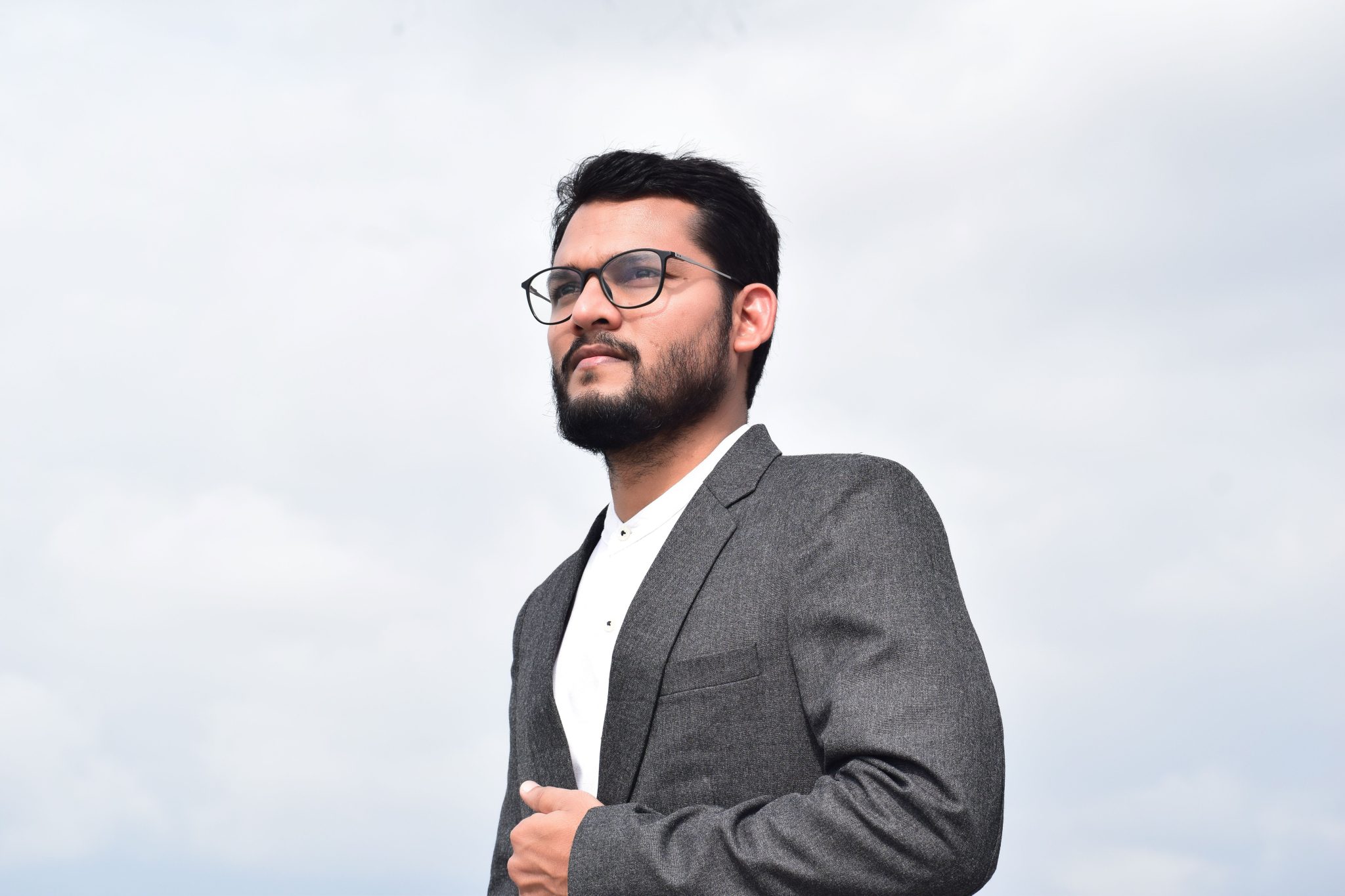 A young businessman wearing glasses confidently poses against a cloudy sky, embodying modern corporate elegance.